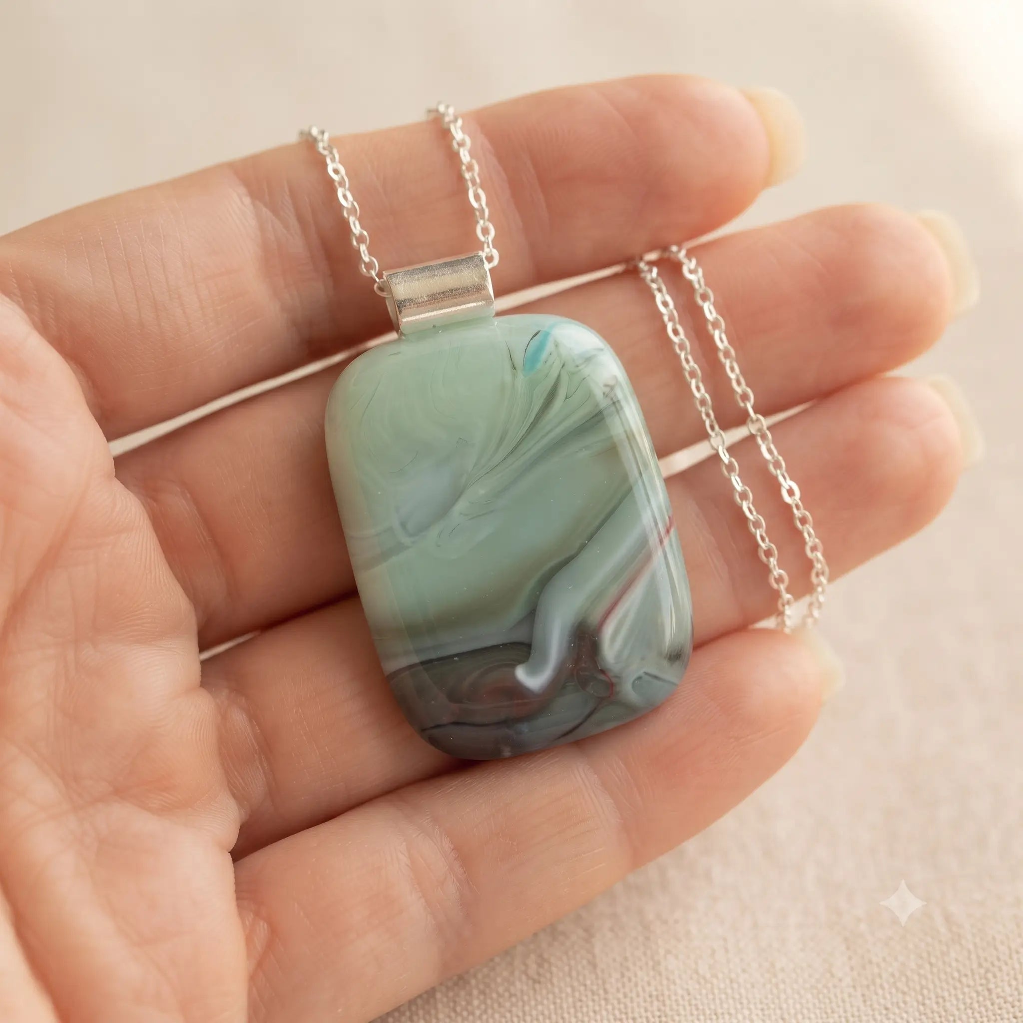 Meadow Mist handmade fused glass pendant in soft sage-mint green with grey and plum-mauve, held in an open palm