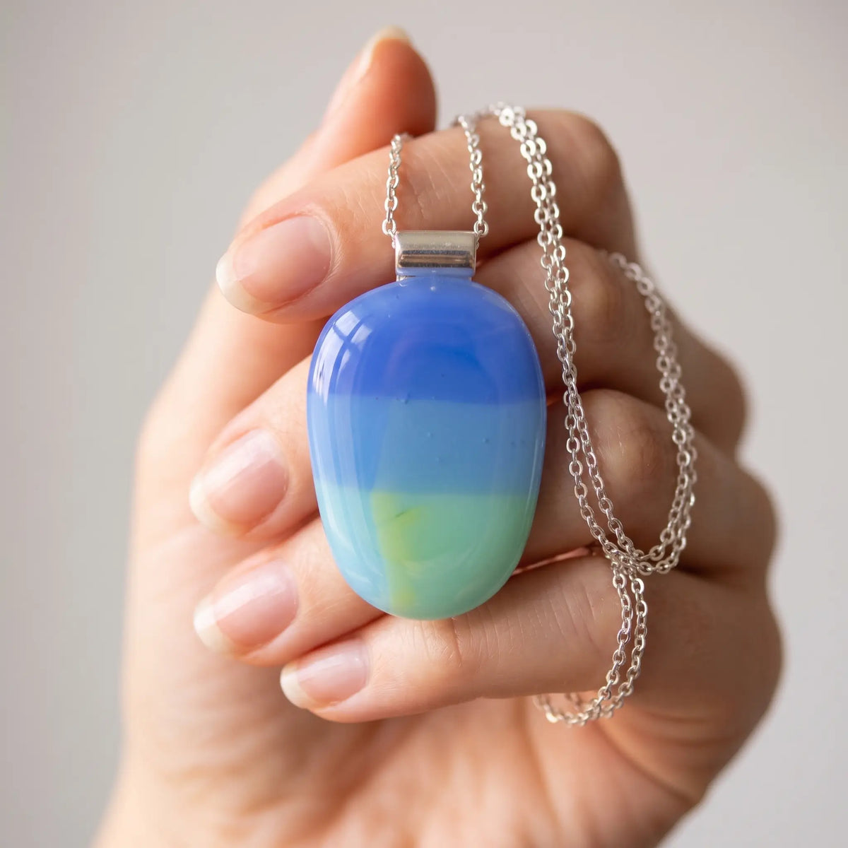 Dayspring handmade fused glass pendant in periwinkle blue, aqua and soft mint, held in an open palm