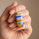 Windbreak handmade fused glass pendant in golden yellow, cobalt and pale sky blue stripes, held in an open palm